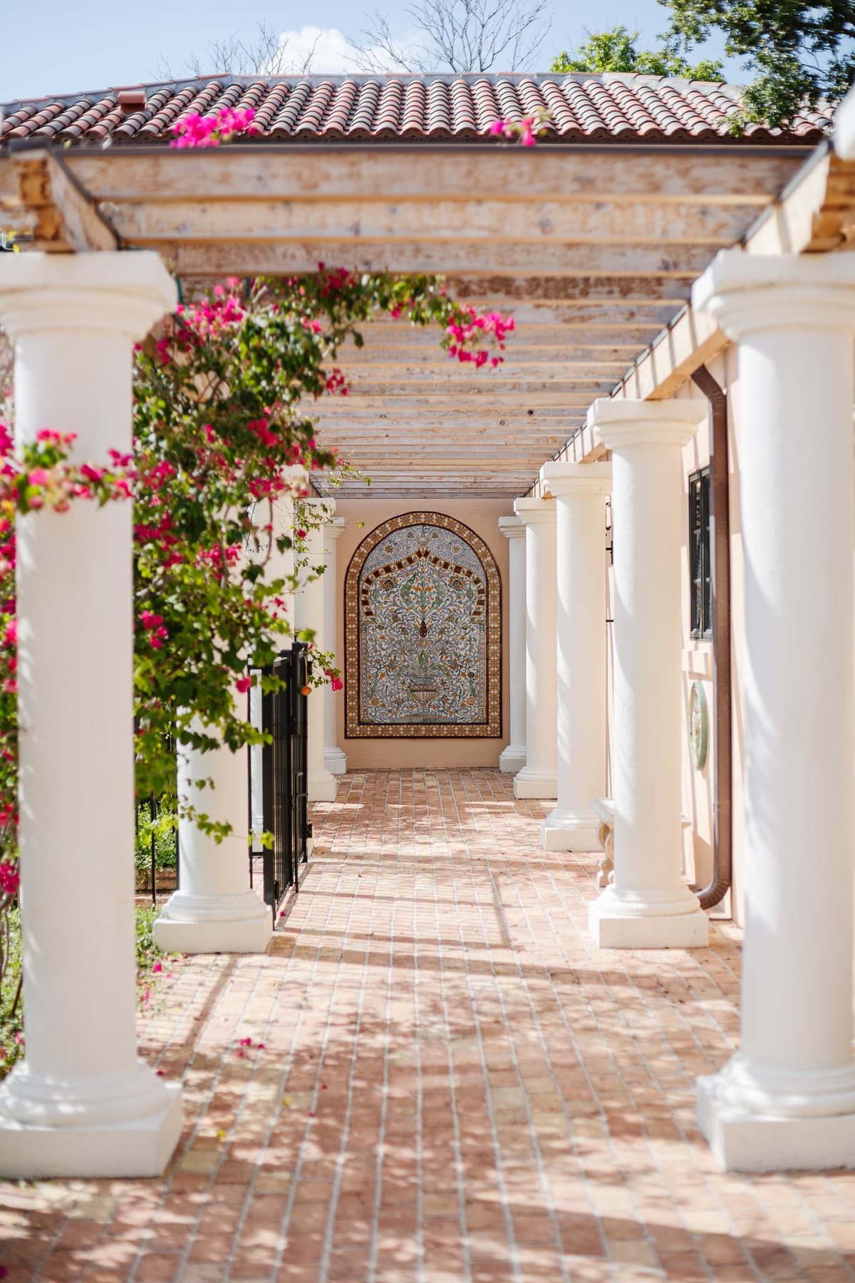 White-columned colonnade with a tiled mural, a wooden pergola, and bougainvillea