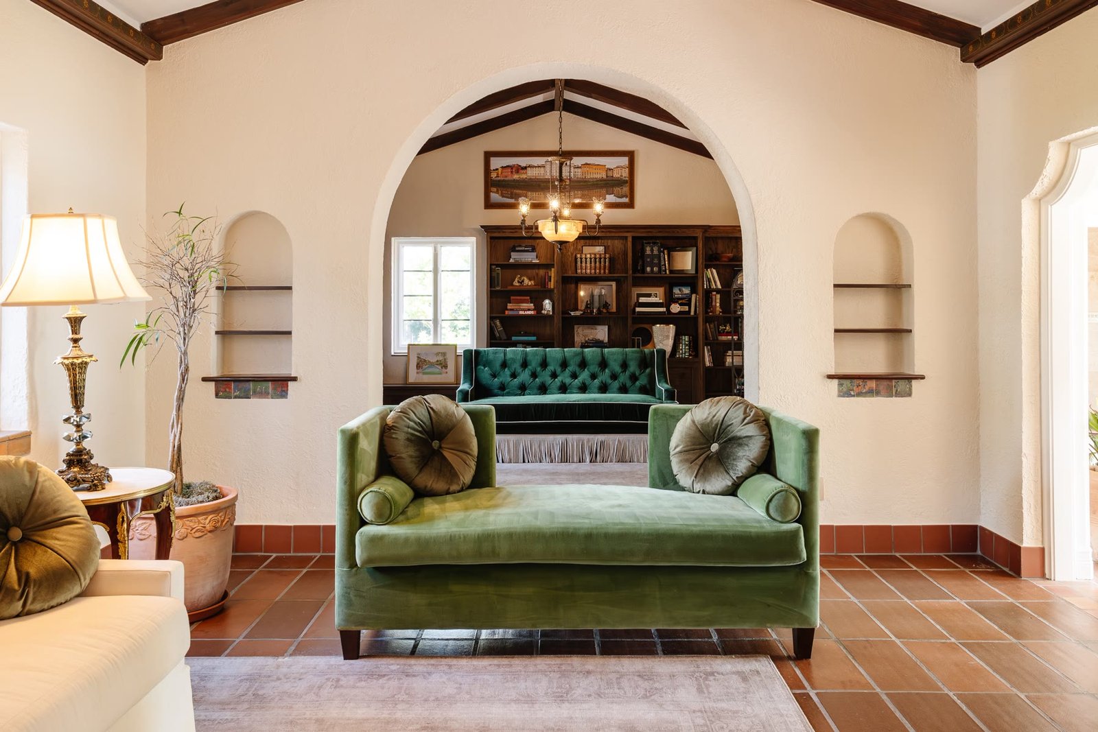 Plaster arch framing the library beyond, with terracotta floors and a green velvet chaise