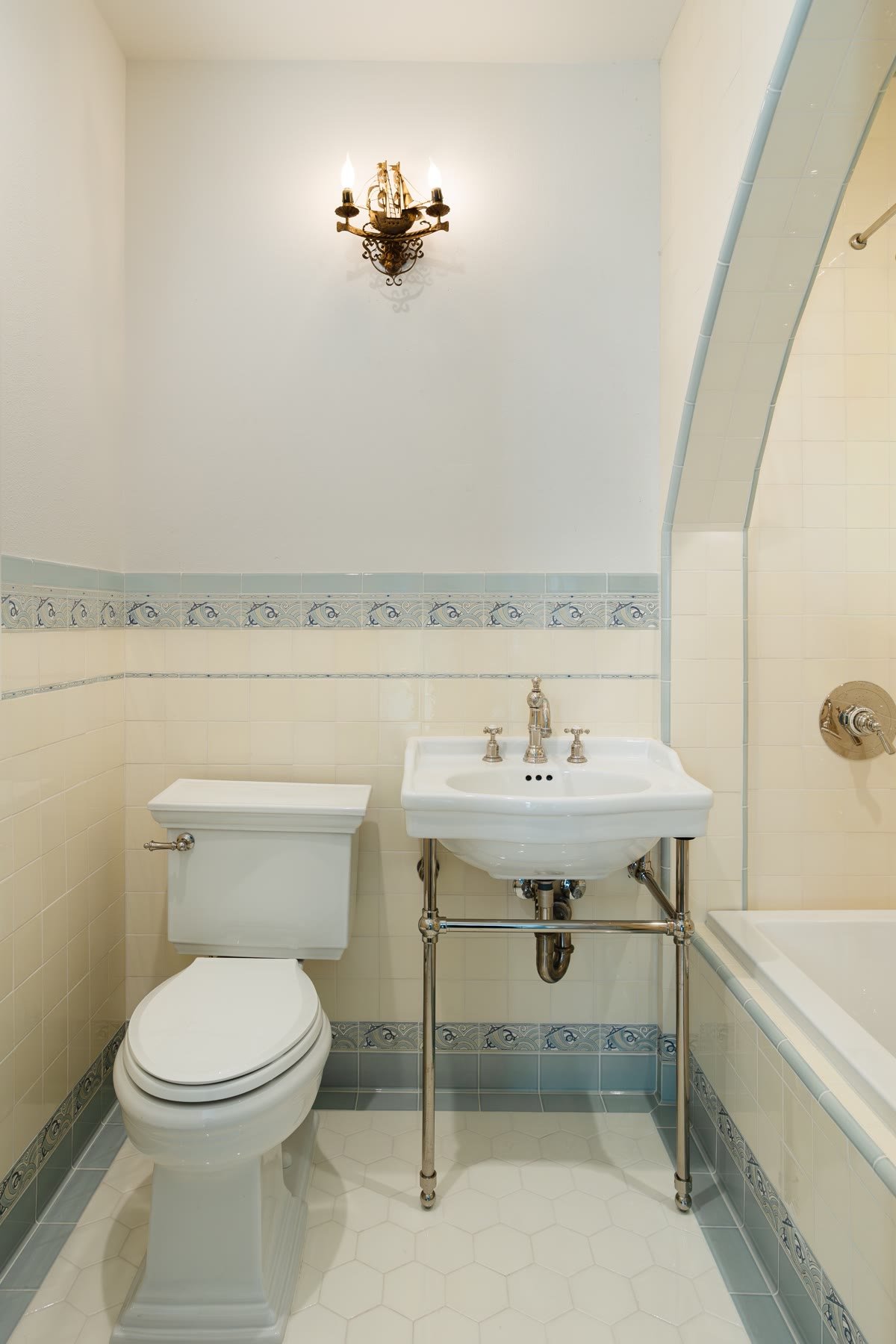 Console sink on chrome legs, cream tile with a blue wave border, and a ship sconce