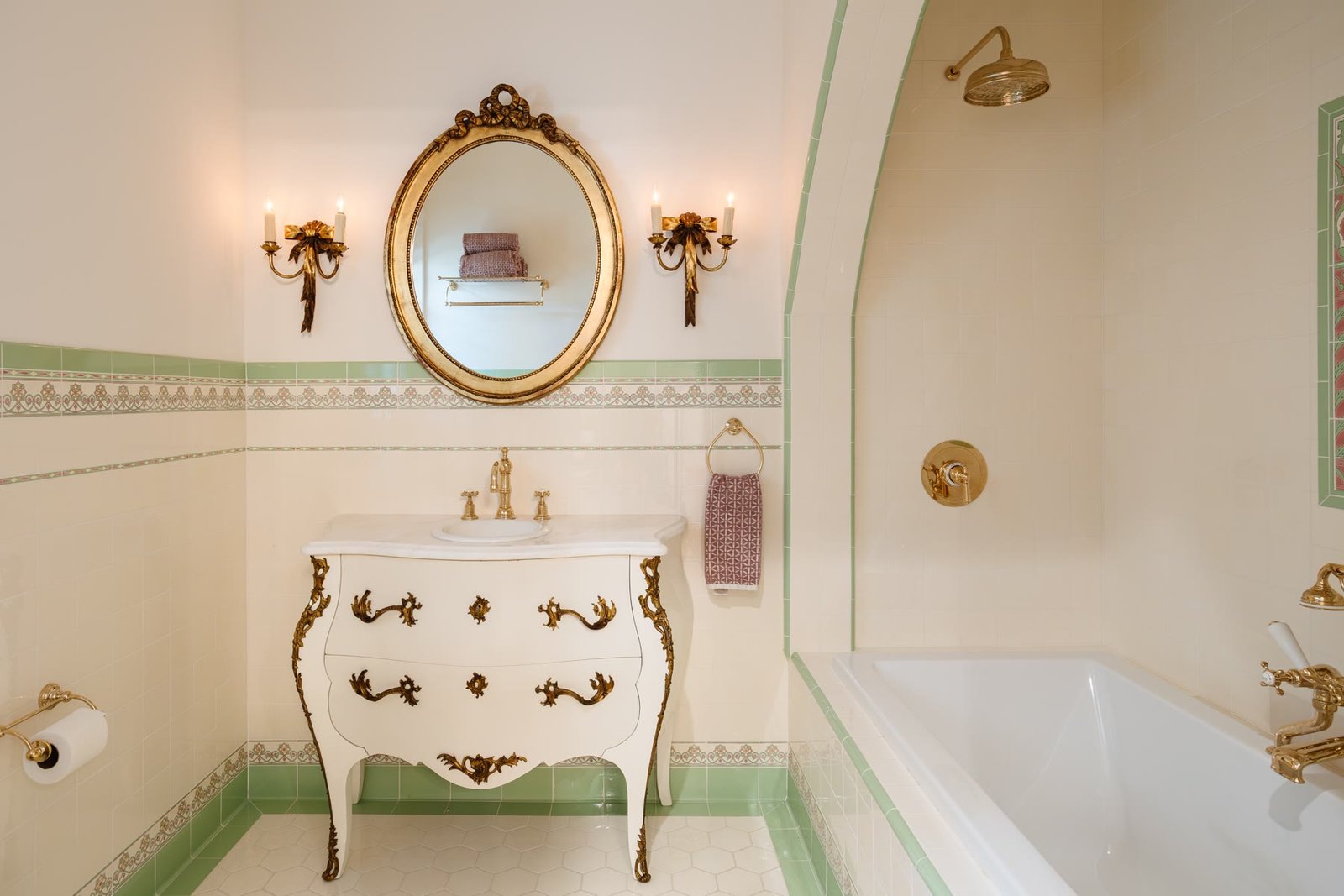 Bath with a French bombe vanity, gilt oval mirror, gold fixtures, and green-bordered tile