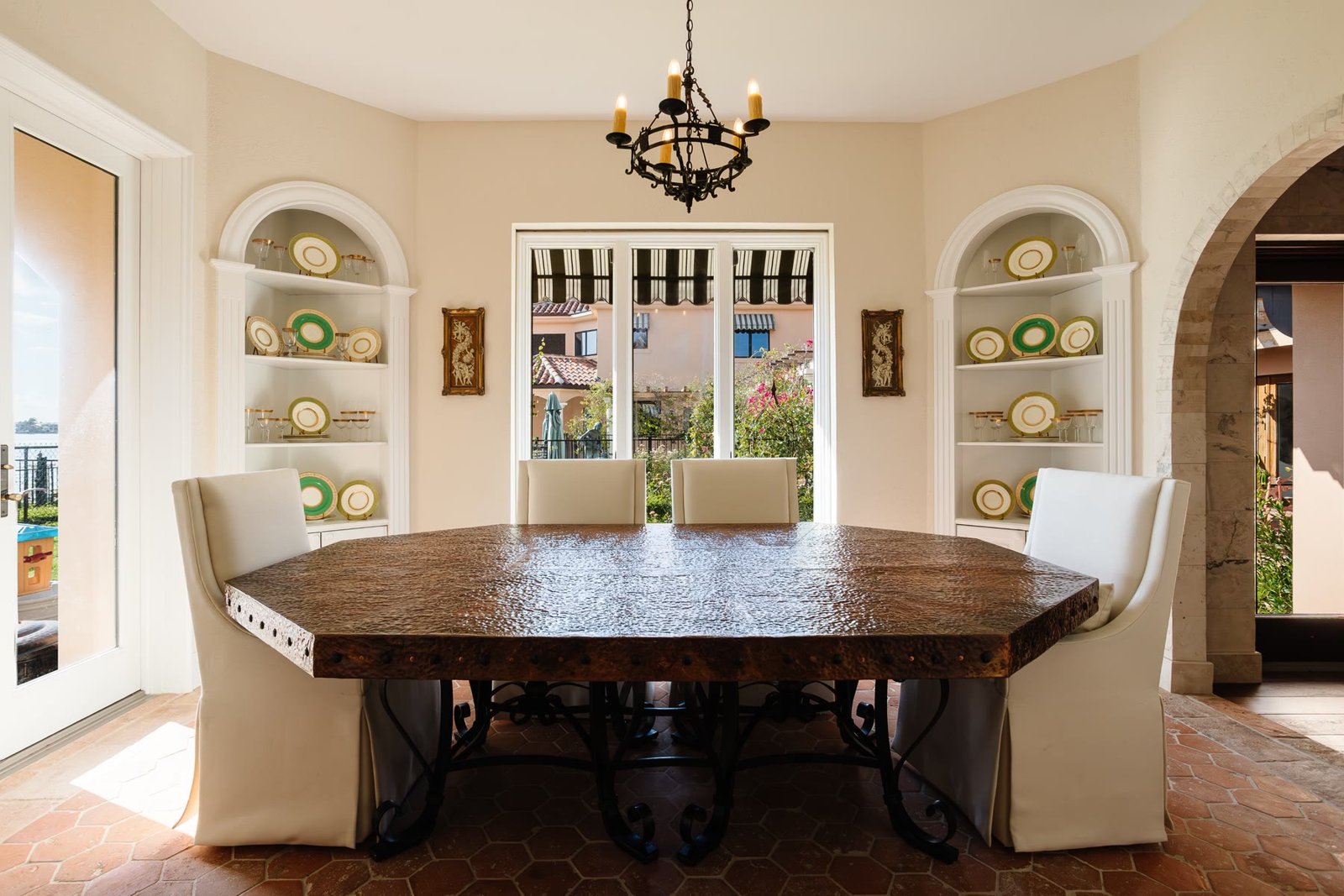 Dining room with a hammered copper table, arched china niches, terracotta hex floor, and a garden view